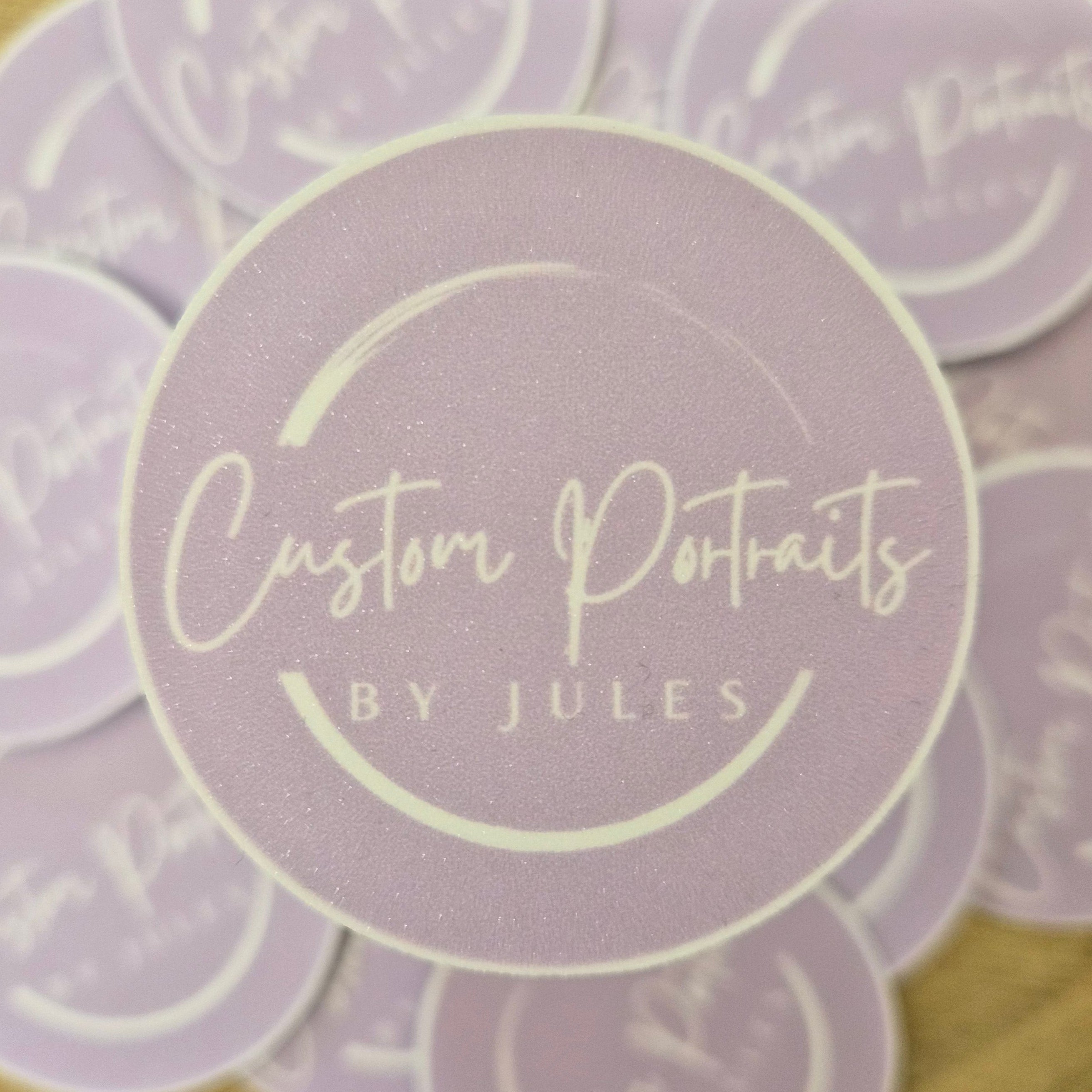 Stack of round purple stickers with 'Custom Portraits by Jules' text on a wooden surface